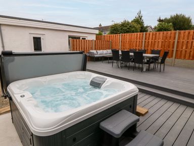 An outdoor patio area with a hot tub a dining table with chairs and a cushioned bench on a wooden deck at Daresfield in Cemaes Bay