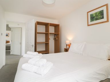 A bedroom with a bed made with white linens towels on the bed a wooden open wardrobe with hangers a lamp on a bedside table and a framed picture on the wall at Harcombe House Bungalow 11 in Chudleigh