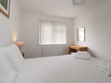 A bedroom with a bed with white linens a nightstand with a lamp a window with vertical blinds and a wooden desk with a mirror at Harcombe House Bungalow 11 in Chudleigh