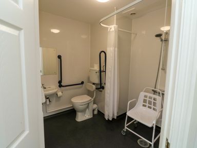 A bathroom with a toilet with support rails a shower area with a shower chair and a sink at Harcombe House Bungalow 11 in Chudleigh