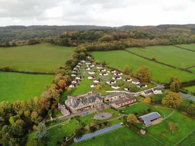 An aerial view of a residential complex with multiple small houses a large building a pond solar panels and surrounding green fields at Harcombe House Bungalow 11 in Chudleigh