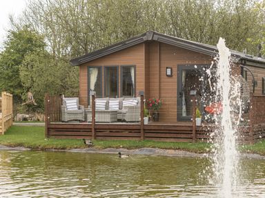 A wooden lodge with outdoor seating on a deck beside a pond with ducks and a water fountain at Loluke Lodge on The Lake 5 Coniston Drive Borwick near Carnforth