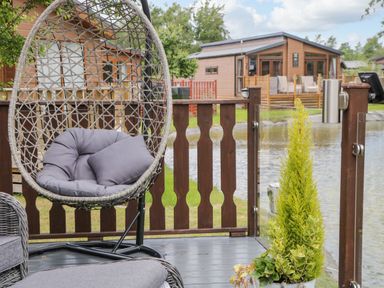An outdoor deck with a hanging chair with cushions a potted plant and a wooden fence overlooking a pond with houses in the background at Loluke Lodge on The Lake 5 Coniston Drive Borwick near Carnforth