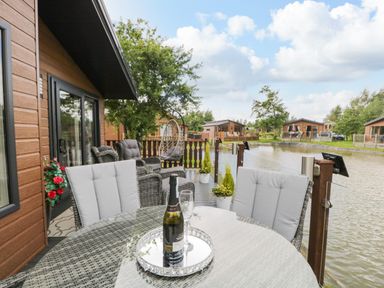 An outdoor deck with a glass table, cushioned wicker chairs, wine bottle and glass overlooking a lake at Loluke Lodge on The Lake 5 Coniston Drive Borwick near Carnforth
