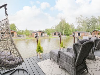A lakeside deck with wicker chairs and hanging chair overlooking water and wooden cabins at Loluke Lodge on The Lake 5 Coniston Drive Borwick near Carnforth