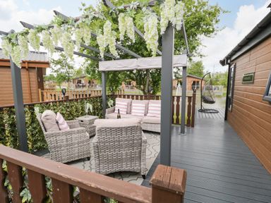 An outdoor deck seating area with wicker furniture under a pergola decorated with hanging flowers at Loluke Lodge on The Lake 5 Coniston Drive Borwick near Carnforth