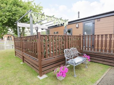A wooden deck with railing and a metal bench with cushions outside a cabin at Loluke Lodge on The Lake 5 Coniston Drive Borwick near Carnforth