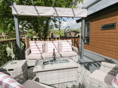 An outdoor seating area with wicker furniture cushions and a tray with a bottle and glasses at Loluke Lodge on The Lake 5 Coniston Drive Borwick near Carnforth