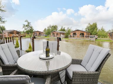 A patio with a glass table and cushioned chairs overlooking a lake and wooden lodges at Loluke Lodge on The Lake 5 Coniston Drive Borwick near Carnforth