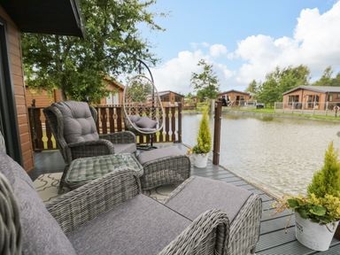 A deck with wicker chairs and a hanging chair next to a lake and wooden lodges at Loluke Lodge on The Lake 5 Coniston Drive Borwick near Carnforth