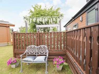 An outdoor seating area with a metal bench with cushions flanked by two flower pots and a wooden fence at Loluke Lodge on The Lake 5 Coniston Drive Borwick near Carnforth