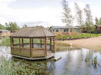 A wooden gazebo on water near a sandy shore with lodges and trees in the background at Loluke Lodge on The Lake 5 Coniston Drive Borwick near Carnforth