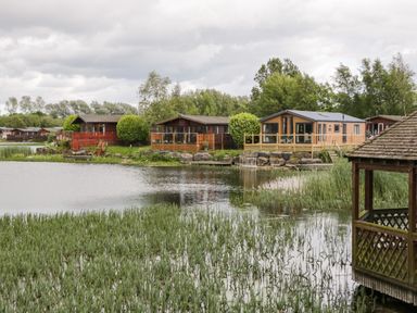 Lake with reeds wooden lodges along the shore and a wooden gazebo at Loluke Lodge on The Lake 5 Coniston Drive Borwick near Carnforth