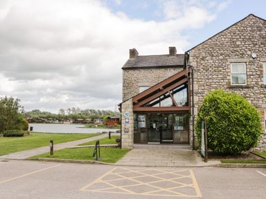 A stone building with glass entrance near a lake and wooden cabins in the background at Loluke Lodge on The Lake 5 Coniston Drive Borwick near Carnforth