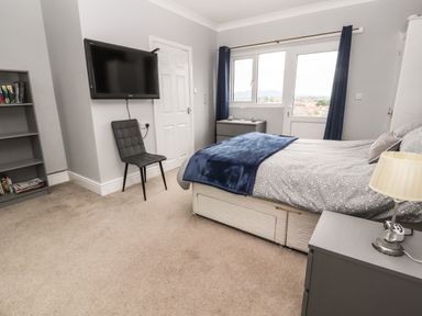 A bedroom with a bed a chair a wall-mounted TV a bookshelf and a window with curtains at Ty Uchaf in Rhyl