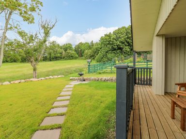 A backyard with a wooden deck a pathway leading to a barbecue grill and trees at The Lodge in Blairgowrie