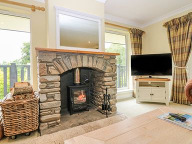 A stone fireplace with a wood burning stove and a basket of firewood beside it next to windows and a television on a stand with curtains in a living room at The Lodge in Blairgowrie