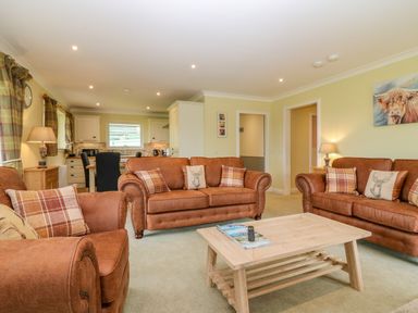 A living room with brown sofas and plaid cushions a wooden coffee table and kitchen in the background at The Lodge in Blairgowrie