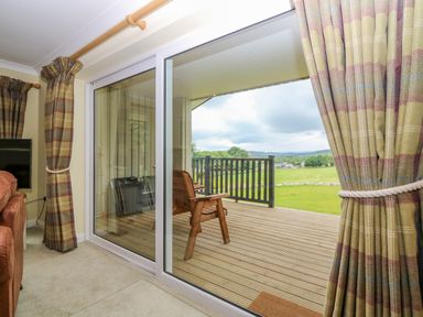 A living room with sliding glass doors leading to a wooden deck with a chair and a view of fields and hills at The Lodge in Blairgowrie