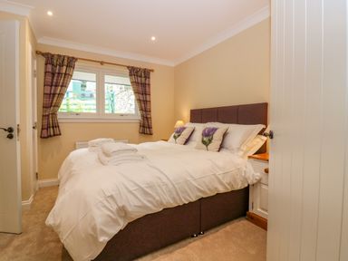 A bedroom with a double bed with white bedding and floral cushions next to a window with plaid curtains at The Lodge in Blairgowrie
