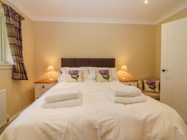A bedroom with a bed with white bedding and floral cushions bedside tables with lamps and a window with plaid curtains at The Lodge in Blairgowrie