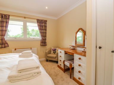 A bedroom with a bed, folded towels, a window with curtains, an armchair, and a wooden dressing table with a mirror at The Lodge in Blairgowrie