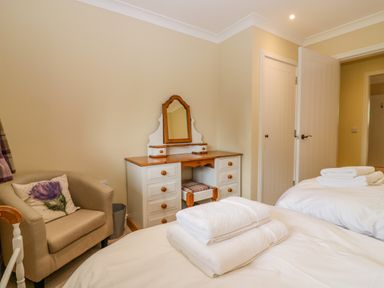 A bedroom with two beds with white linens and folded towels on top a beige armchair with a flower cushion and a wooden dressing table with a mirror at The Lodge in Blairgowrie