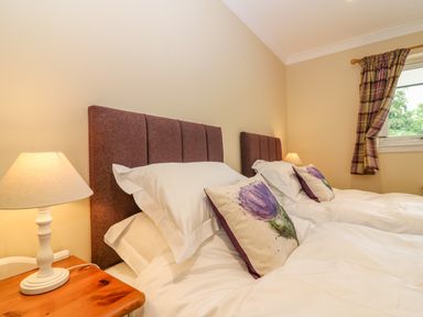 A bedroom with two single beds side by side with purple headboards and floral cushions two bedside tables each with a lamp and a window with plaid curtains at The Lodge in Blairgowrie