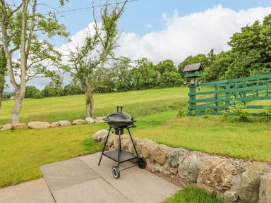 A charcoal barbecue on a stone patio next to a low stone wall with a green wooden fence and bird feeder in a grassy garden at The Lodge in Blairgowrie