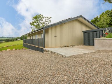 A gravel driveway leading to a one-story wooden house with a railing and a grassy field in the background at The Lodge in Blairgowrie