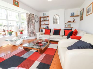A living room with white sofas red and black cushions a patterned rug glass coffee tables a bookshelf and large windows at 2 Old Talbot Cottages Lostwithiel