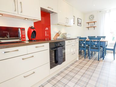 A kitchen with white cabinets red microwave and coffee machine blue dining table with chairs and clock on wall at 2 Old Talbot Cottages in Lostwithiel