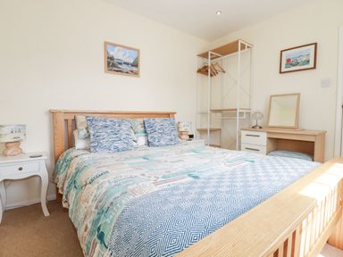 A bedroom with a wooden bed frame and patterned bedding a white bedside table with a lamp an open wardrobe and a dressing table with a mirror at 2 Old Talbot Cottages in Lostwithiel