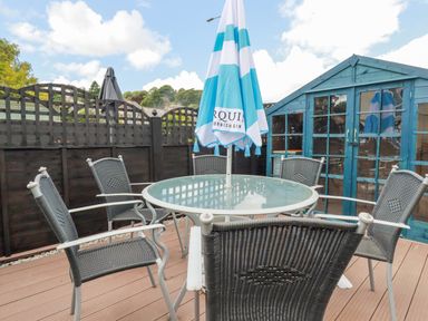 An outdoor patio with a glass table surrounded by six chairs and a blue striped umbrella near a blue wooden shed at 2 Old Talbot Cottages in Lostwithiel