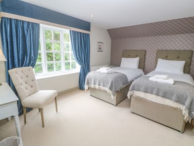 A bedroom with two single beds a window with blue curtains and a beige chair at Bodmin Country House Lanivet