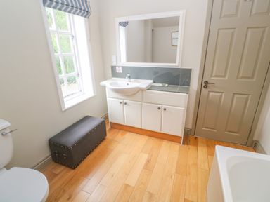 A bathroom with a sink and mirror next to a window with a striped blind and a dark storage bench on a wooden floor at Bodmin Country House in Lanivet
