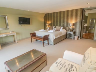 A bedroom with a bed, leather bench, TV on the wall, desk with mirror, chair, and doorway to a bathroom at Bodmin Country House in Lanivet