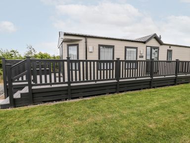 An exterior view of a beige mobile home with black framed windows and black railing on a deck with grass in the foreground at Lazy Daze in Carnaby near Bridlington