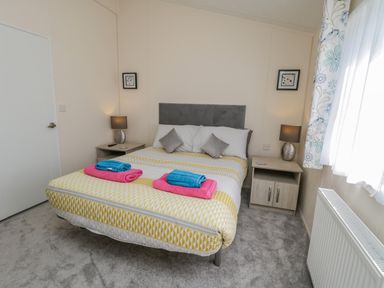 A bedroom with a bed with yellow patterned bedding and folded towels on top two bedside tables with lamps and framed pictures on the walls at Brighter Daze in Carnaby near Bridlington