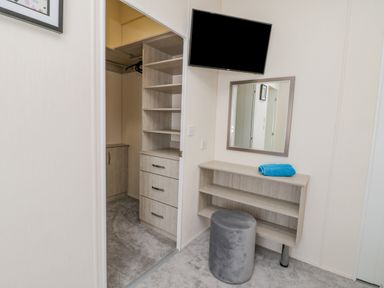 A room with a wall-mounted television a mirror a small desk with shelves a round stool and an open closet with drawers and shelves at Brighter Daze in Carnaby near Bridlington
