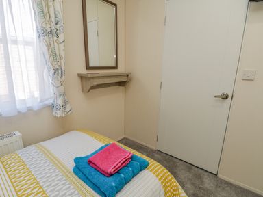 A bedroom with a bed covered by a patterned duvet and towels on top a wall-mounted shelf with a mirror and a closed white door at Brighter Daze Carnaby near Bridlington