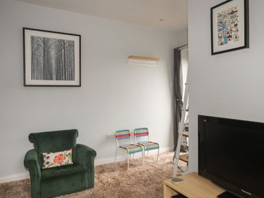 A room with a green armchair, two colorful chairs, a wooden shelf, framed pictures on the wall, and a television on a wooden stand at The Old Coal Yard in Cemaes Bay