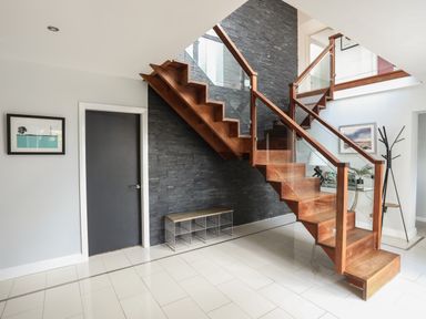 A wooden staircase with glass panels and a black stone wall behind it in a tiled hallway with a black door and a shoe rack at The Old Coal Yard in Cemaes Bay
