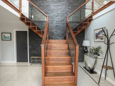 A wooden staircase with glass panels on a black stone wall with a black door and a console table with a lamp and plant at The Old Coal Yard in Cemaes Bay
