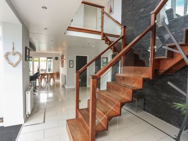 An interior view of a hallway with a wooden staircase and glass railing leading to an upper floor and a dining area with chairs and a table visible in the background at The Old Coal Yard in Cemaes Bay