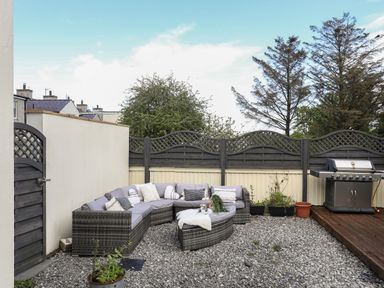 An outdoor patio with a wicker sectional sofa and cushions a grill and a gravel ground at The Old Coal Yard in Cemaes Bay