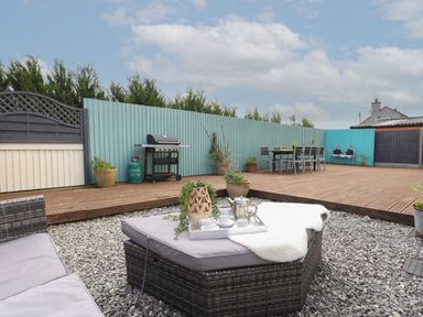 An outdoor patio area with a barbecue grill a dining table with chairs and cushioned seating on gravel and wooden decking at The Old Coal Yard in Cemaes Bay