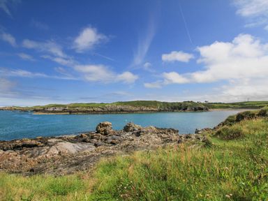 A coastal scene with rocky shore grass and an island in the distance under a blue sky at The Old Coal Yard in Cemaes Bay