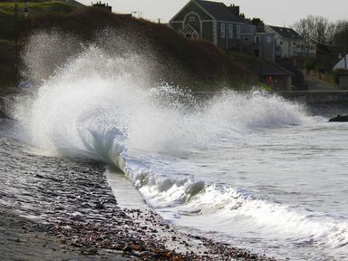 Waves crashing against a seawall with houses on a hill in the background at The Old Coal Yard in Cemaes Bay