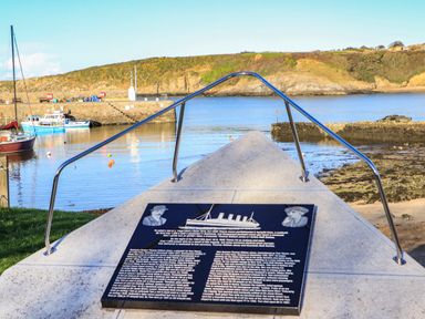 A memorial plaque on a concrete platform by a harbor with boats and hills in the background at The Old Coal Yard in Cemaes Bay